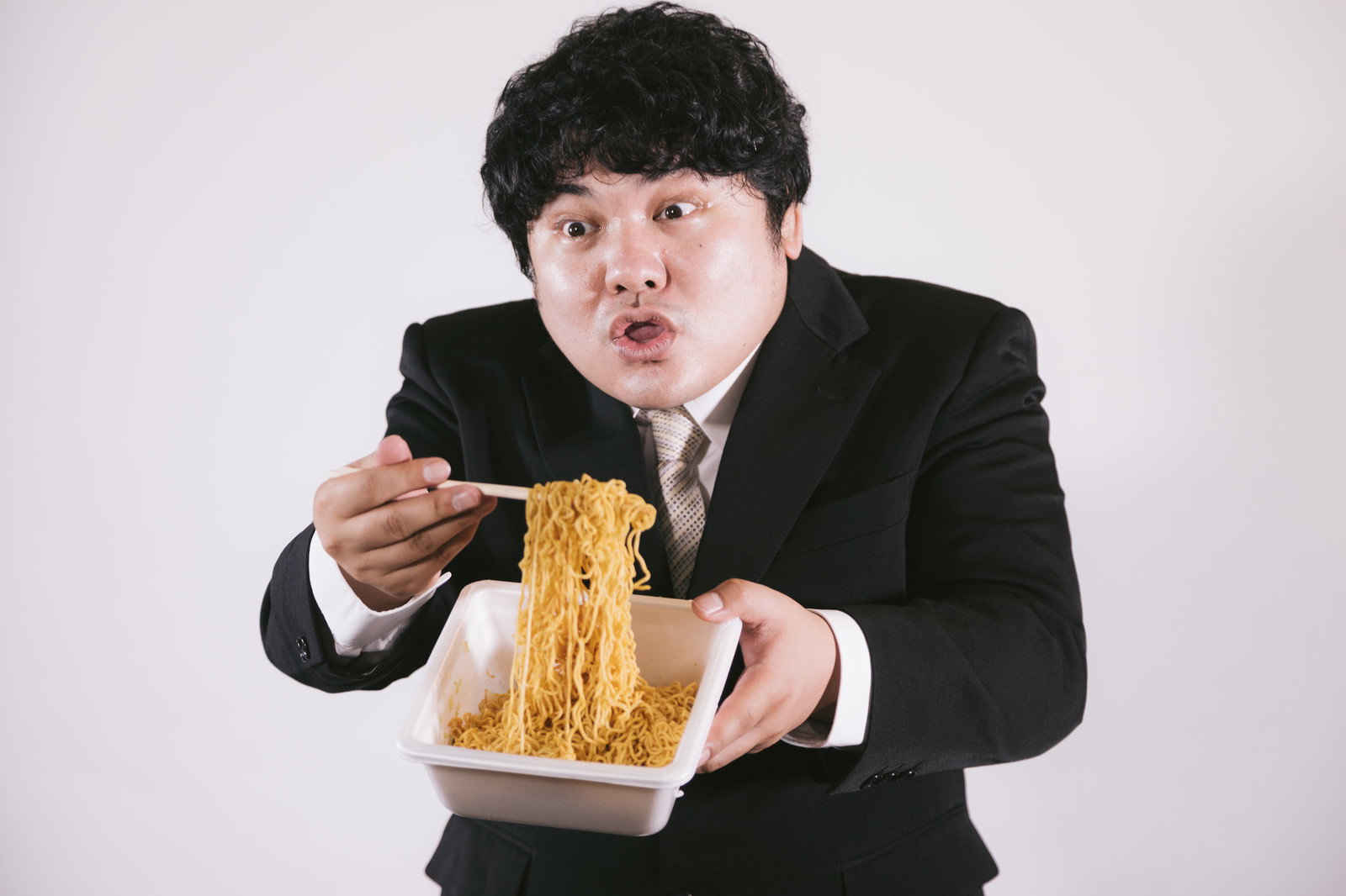黒いスーツを着た男性がカップ焼きそばを箸で持ち上げ、驚いた表情を見せている［モデル：段田隼人］