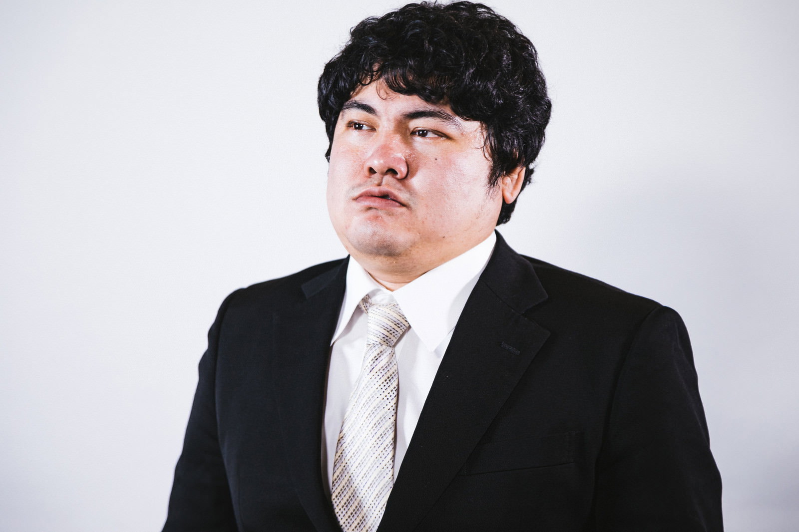 黒いスーツで不満そうな表情をする男性の正面顔写真［モデル：段田隼人］