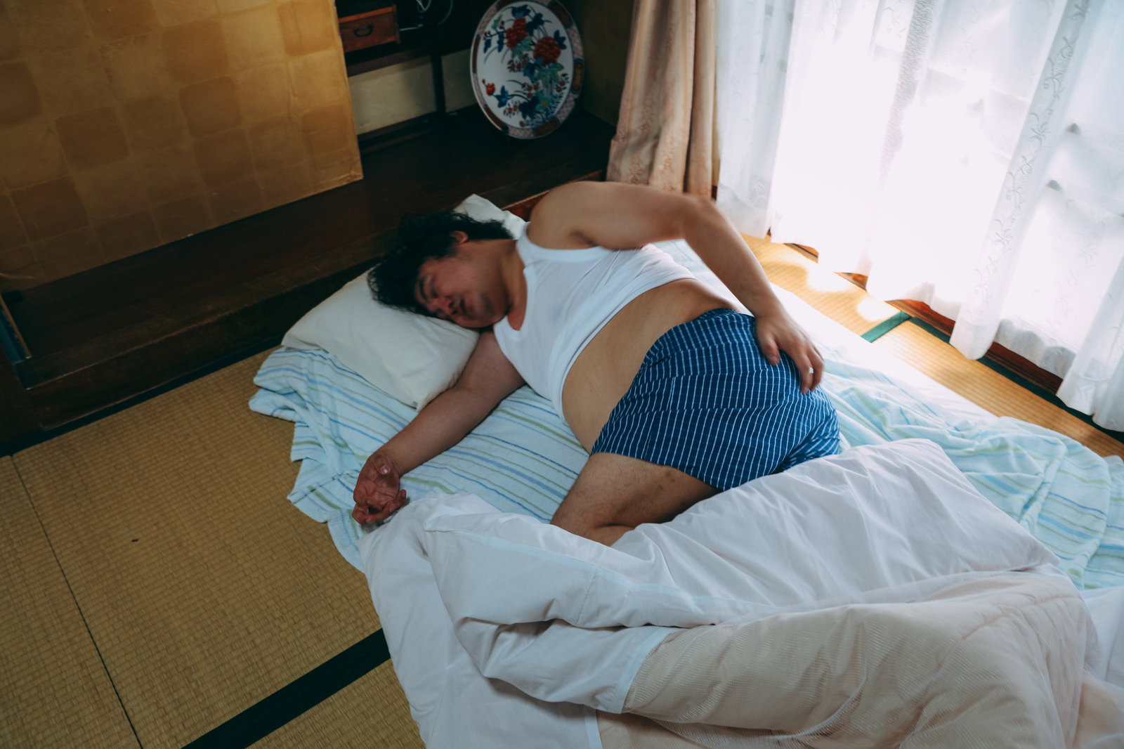 昼間に布団の上で寝ている肥満体の男性［モデル：段田隼人］