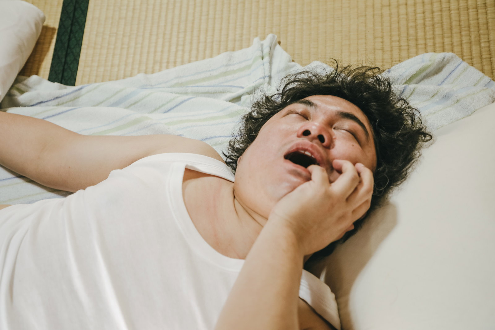 畳の上で口を開けたまま熟睡する、タンクトップ姿の男性［モデル：段田隼人］