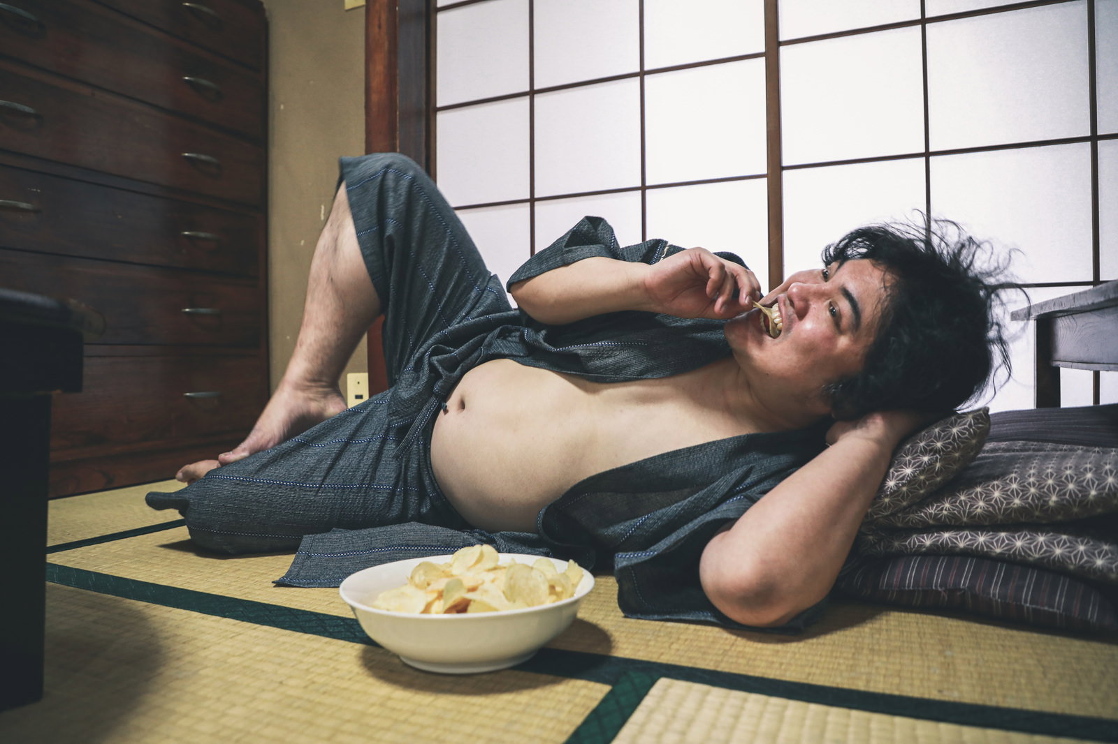 腹を出してポテチを食べながら和室の畳で寝転ぶ男性［モデル：段田隼人］