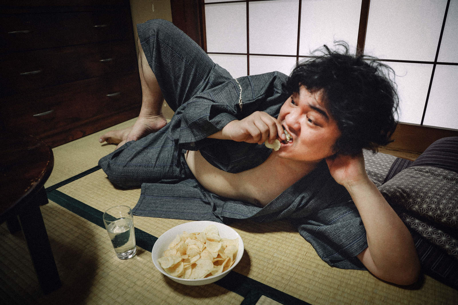 畳の上でコップ酒とポテチを楽しみながら横たわる男性［モデル：段田隼人］