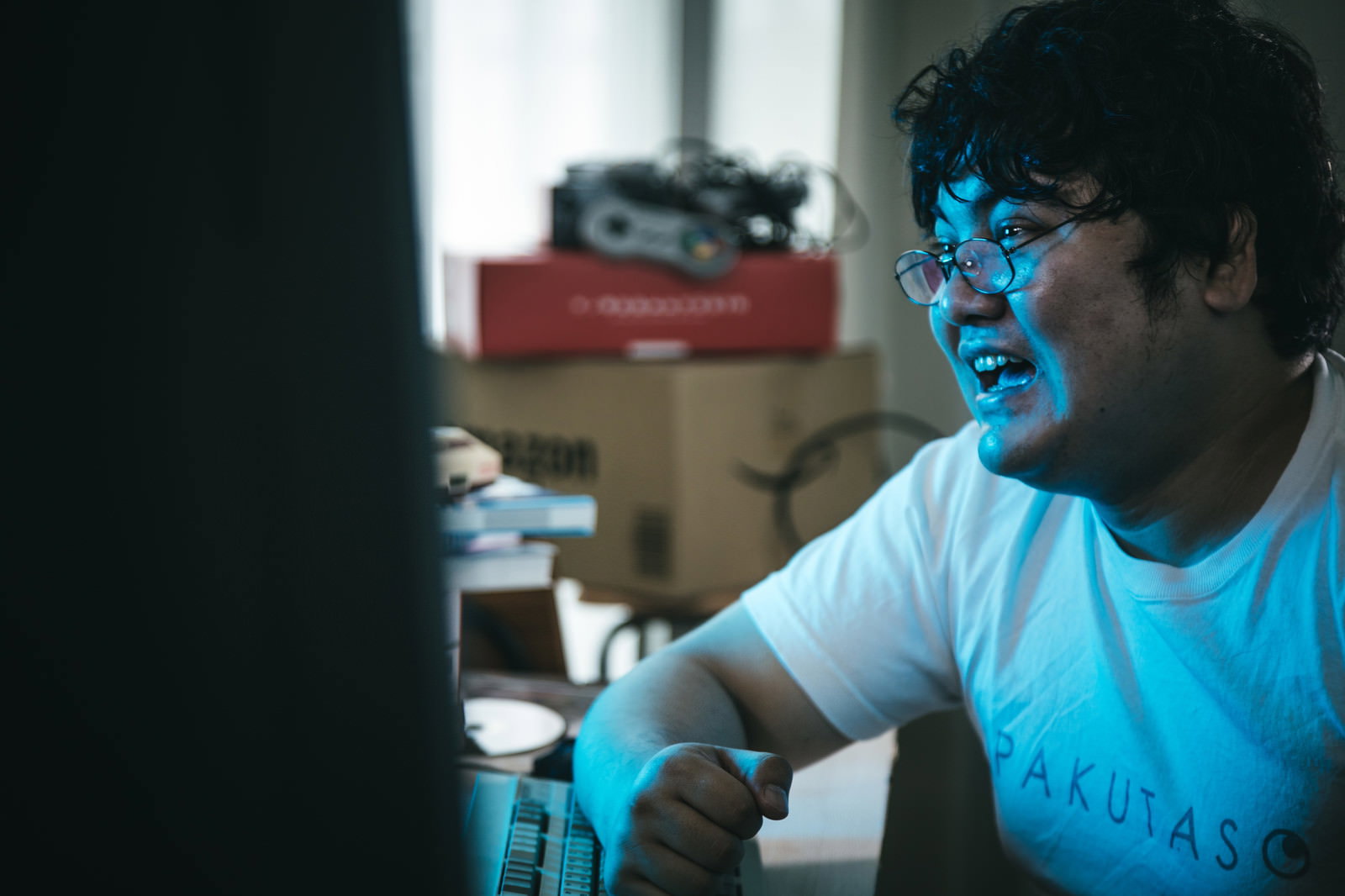 デスクでキーボード方向に身を乗り出し、激しい表情でポーズを取る眼鏡の男性［モデル：段田隼人］