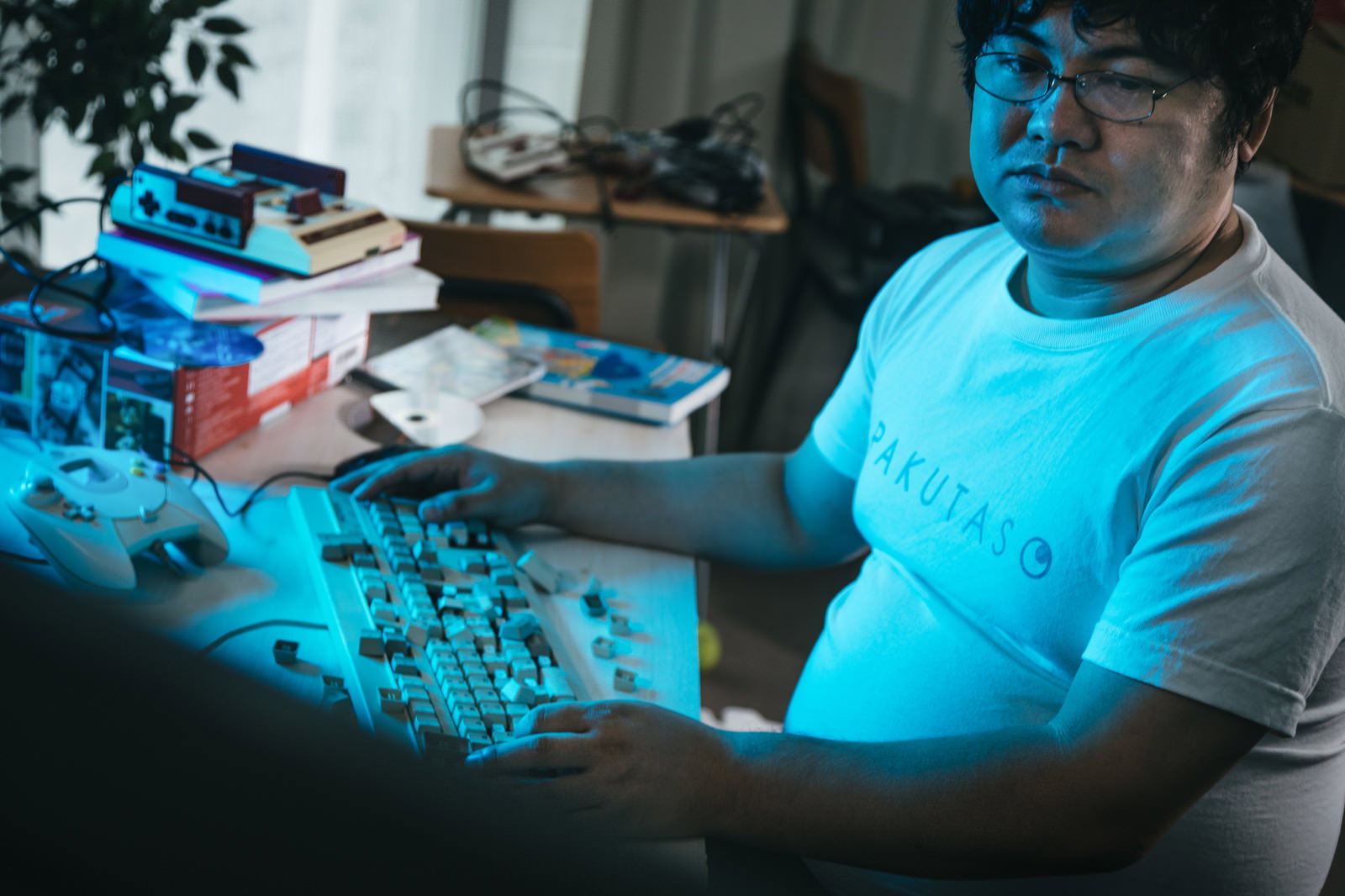 暗い部屋でモニターの青い光に照らされながらキーボードを操作する眼鏡の男性［モデル：段田隼人］