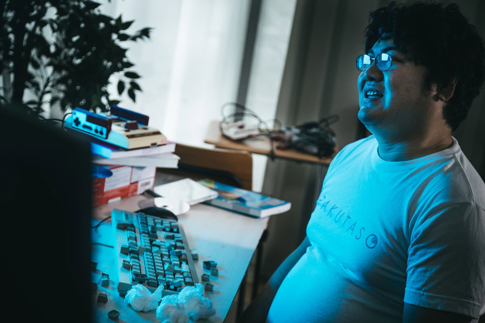 ゲーミングチェアに座り、デスクの前で上を向く疲れた男性。青いネオン照明が照射された室内［モデル：段田隼人］