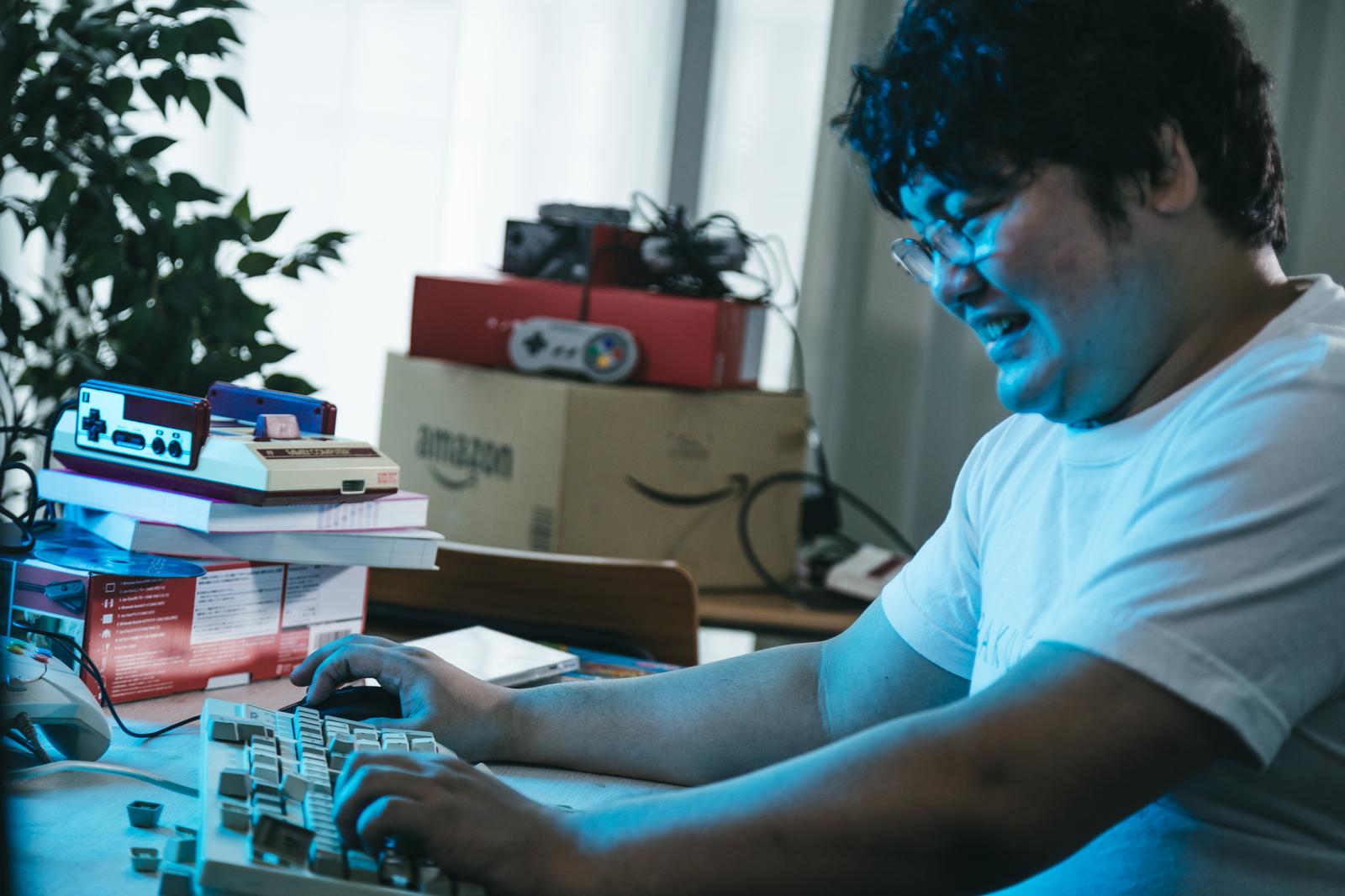 青い光の中でキーボードを叩いている男性［モデル：段田隼人］