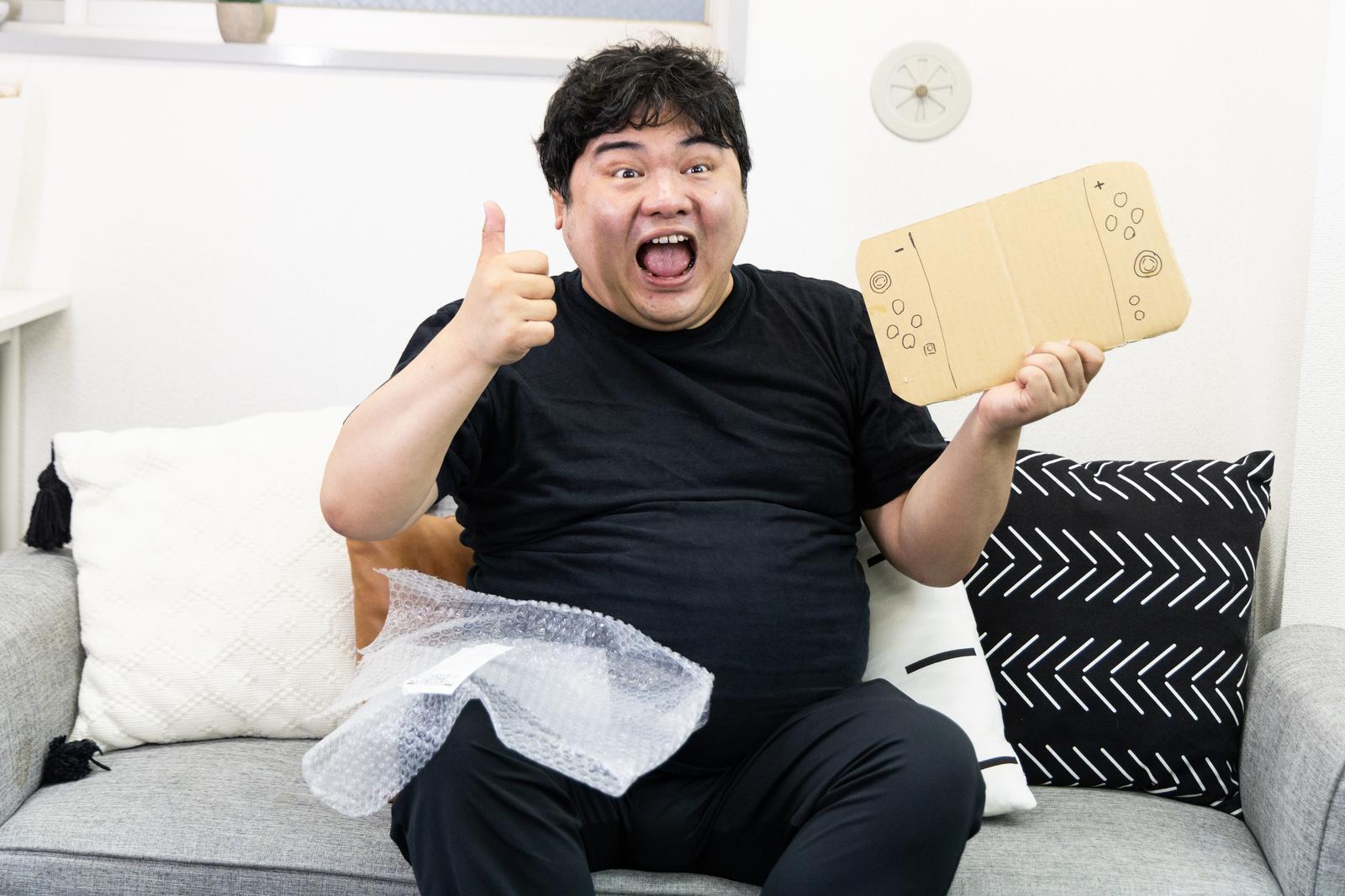 Man in black t-shirt holding cardboard game console, giving thumbs up and smiling