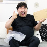 黒Tシャツ姿で段ボール製ゲーム機を手に親指を立てる男性の写真