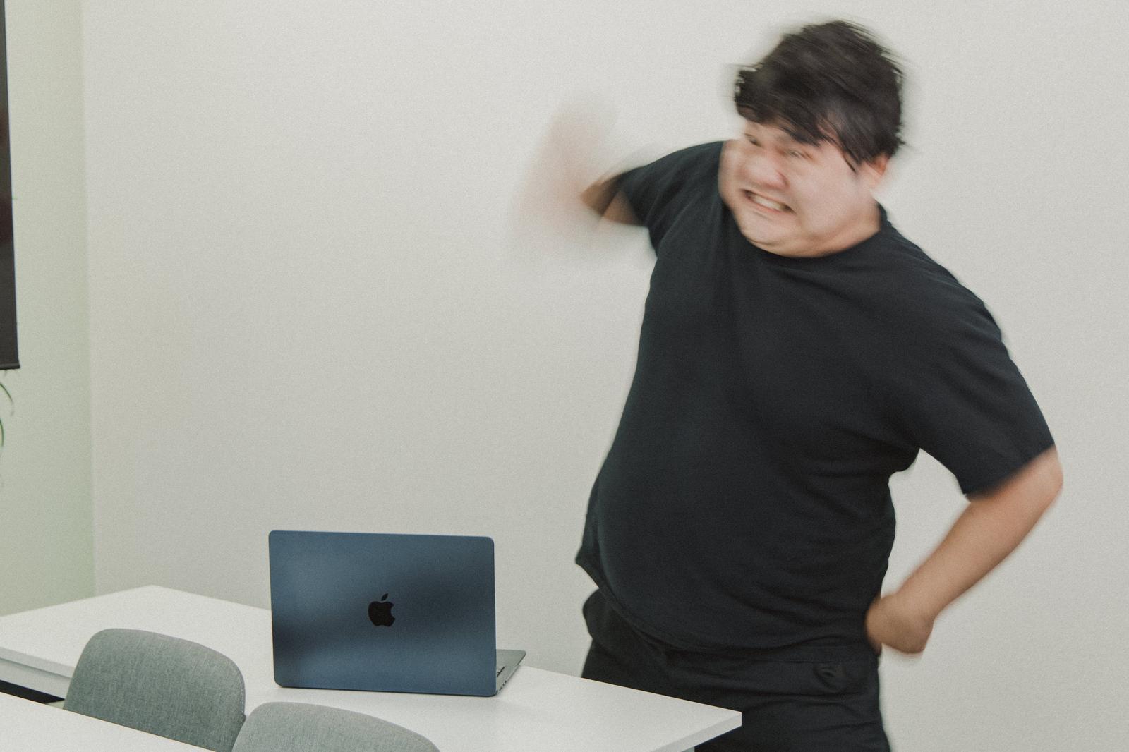 白いデスクでノートパソコンに向かい激しく怒る黒Tシャツの男性［モデル：段田隼人］