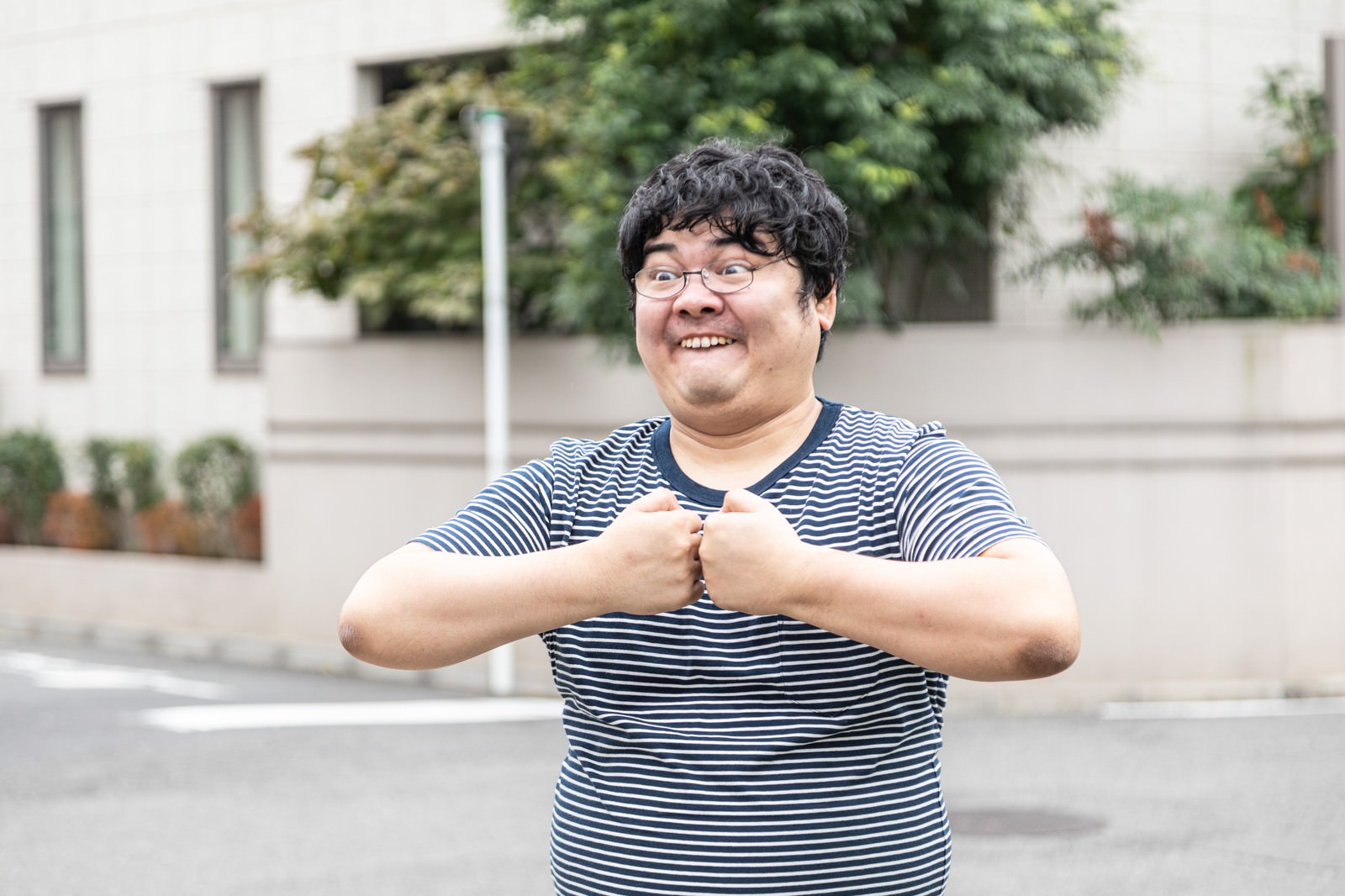 ストライプシャツを着たメガネをかけた男性が両手で拳を作り力を込めている［モデル：段田隼人］