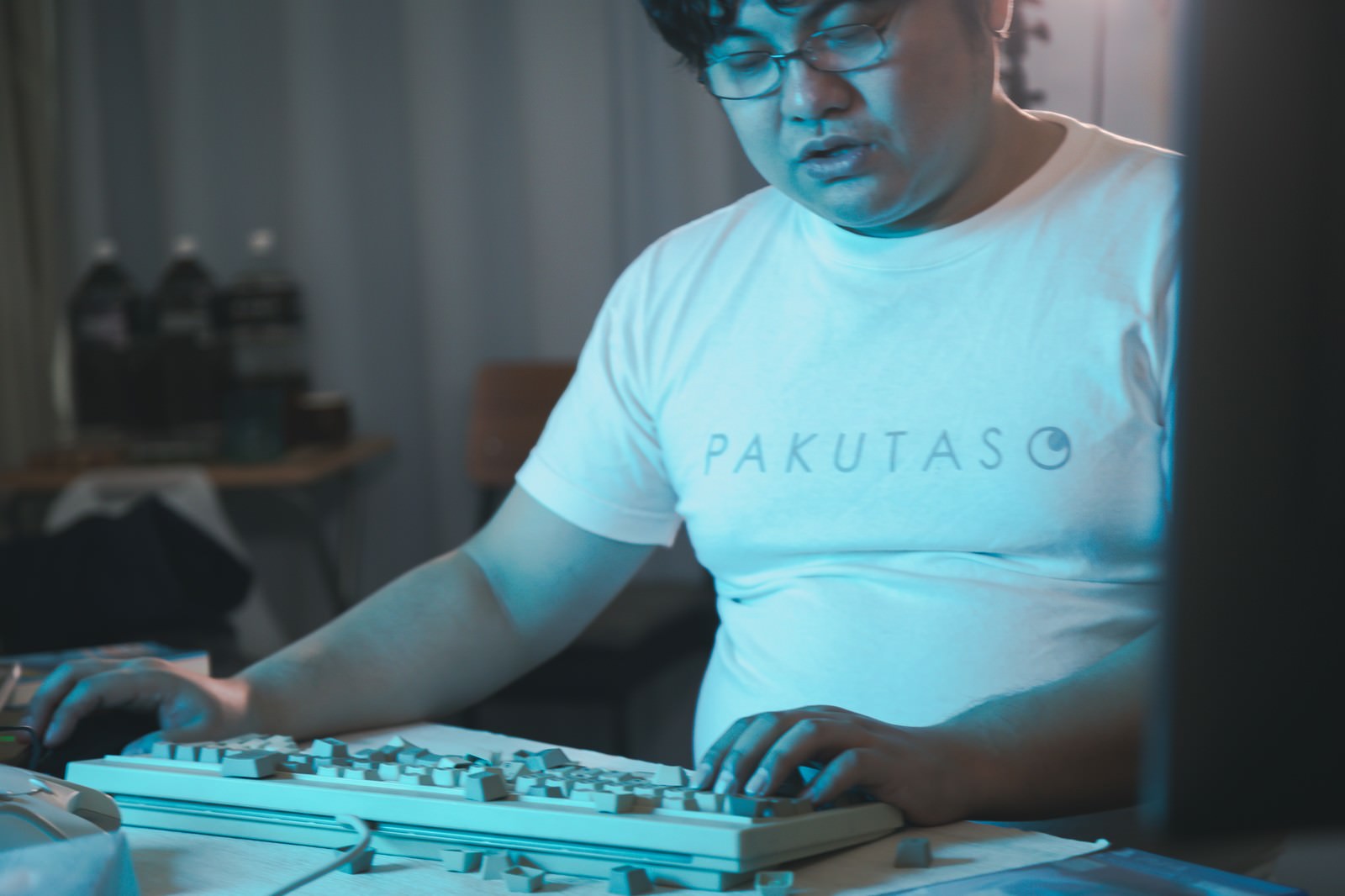 青いライトの中でキーボードに手を置く白いTシャツの男性［モデル：段田隼人］