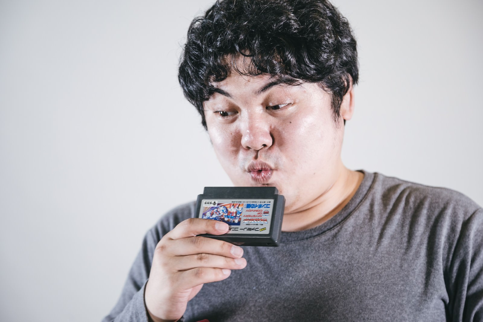 ファミコンカセットに息を吹きかけるグレーのTシャツを着た男性［モデル：段田隼人］