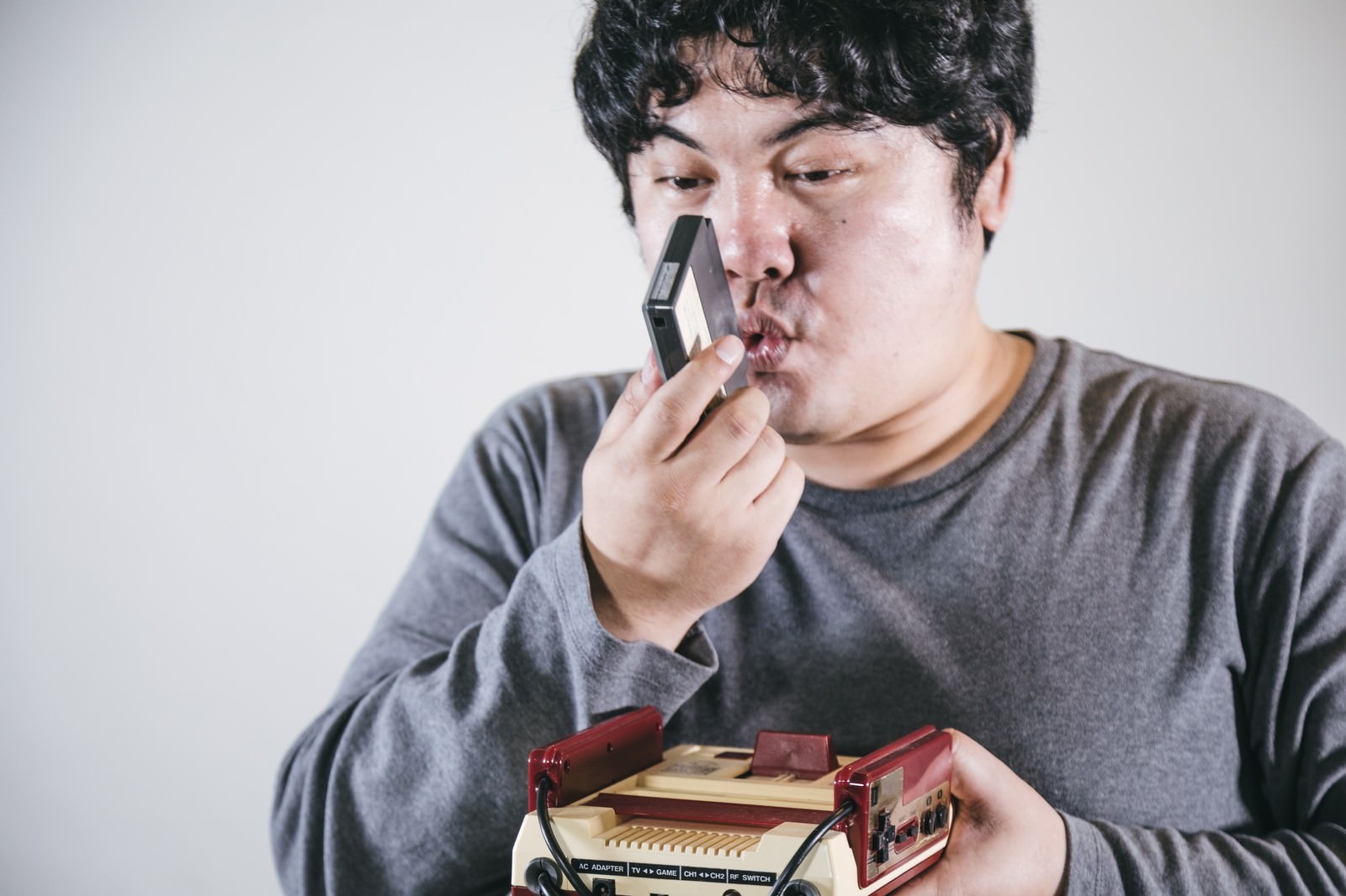 グレーのTシャツを着た男性がゲームカセットに息を吹きかけている様子［モデル：段田隼人］