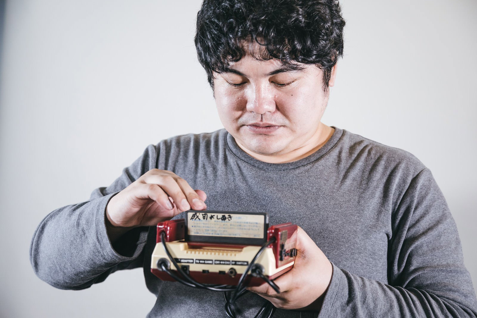 グレーのトレーナーを着た男性がファミコンカセットを本体に差し込んでいる様子［モデル：段田隼人］