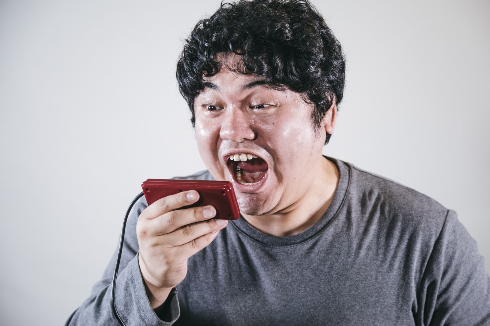 赤いゲームコントローラーを持ち大声で叫ぶグレーTシャツの男性［モデル：段田隼人］