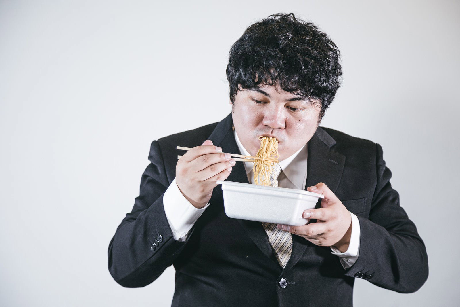 黒いスーツを着た男性がカップ焼きそばを箸で食べている［モデル：段田隼人］