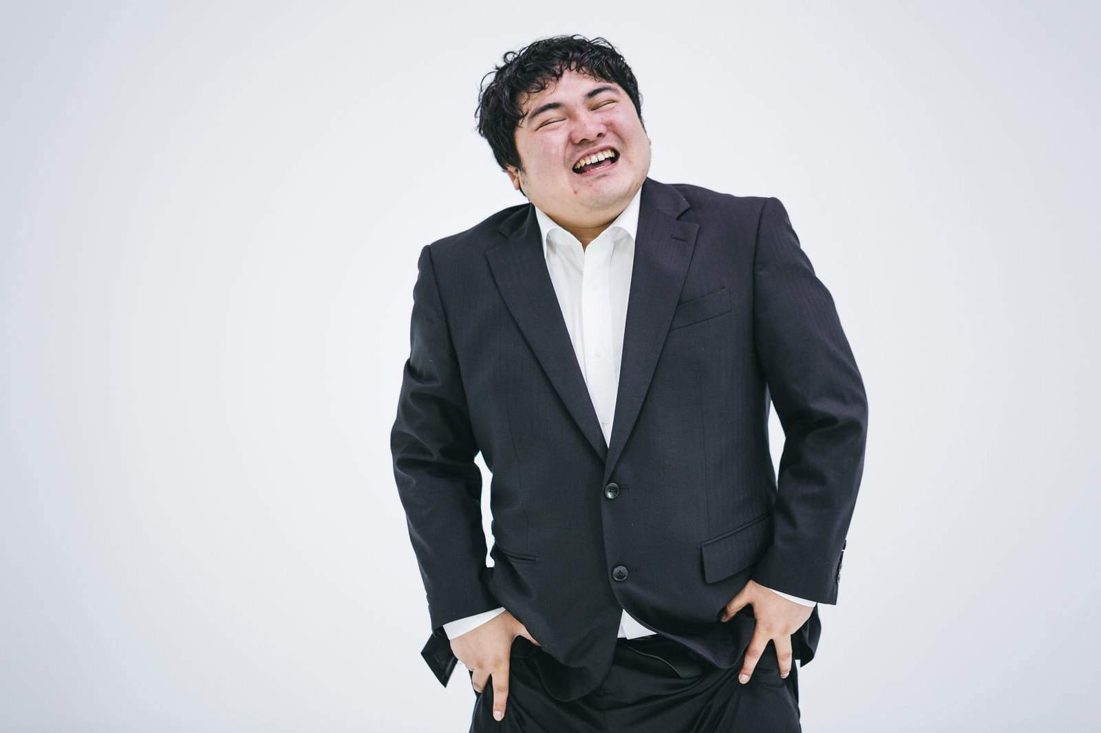 腰に手を当てて体を傾け、ニヤニヤ笑ってごまかすスーツ姿の男性［モデル：段田隼人］