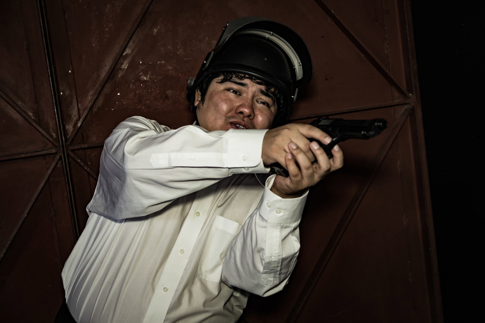 物陰に隠れて拳銃を両手で構える白いシャツの男性［モデル：段田隼人］