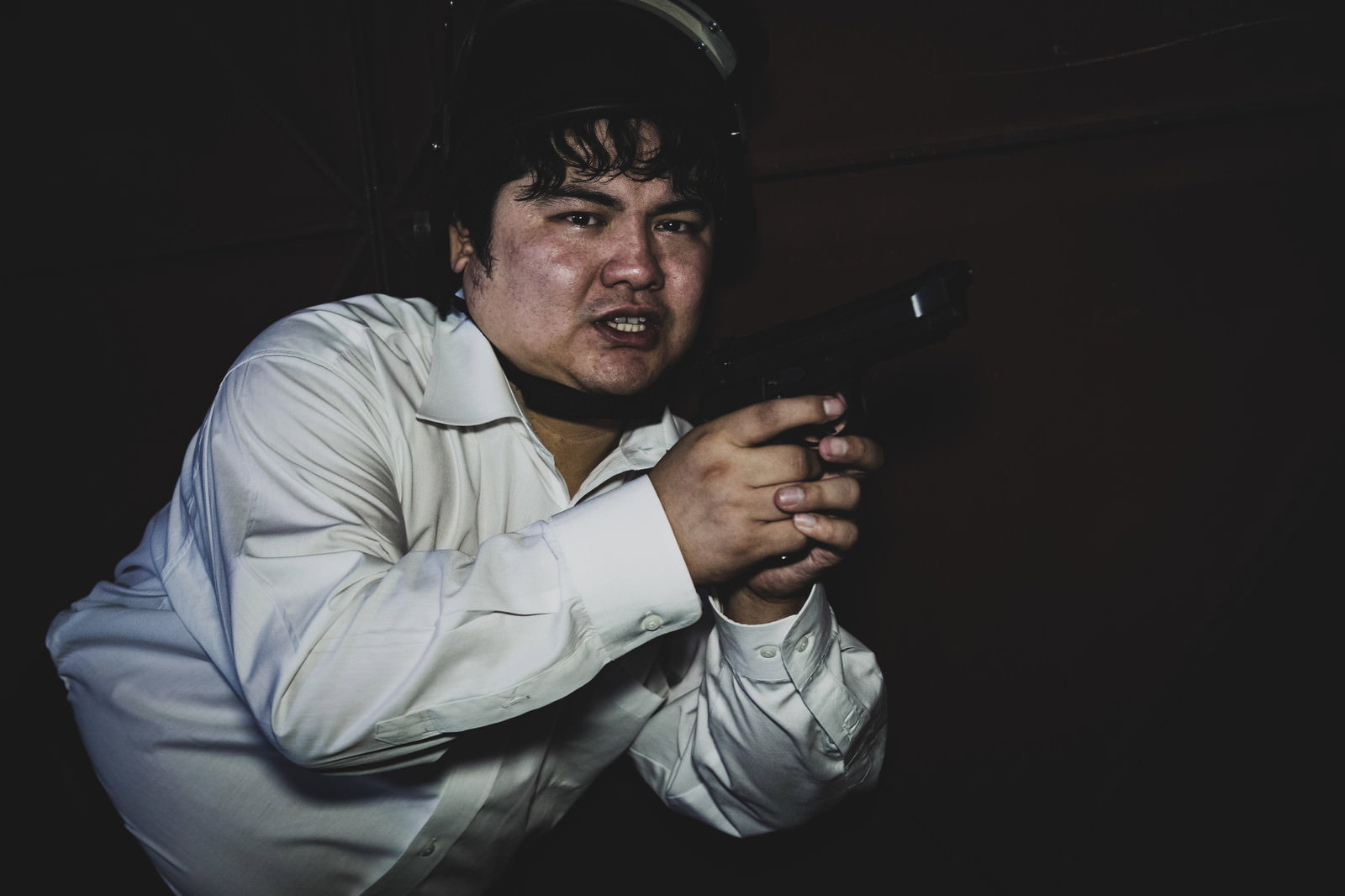暗い室内で拳銃を両手で構えて警戒する白いシャツを着た男性［モデル：段田隼人］