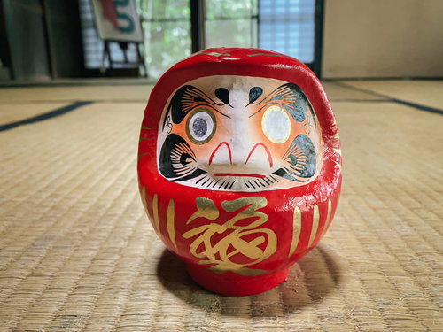和室の畳に置かれた片目のままの赤い達磨さんの祈願置物