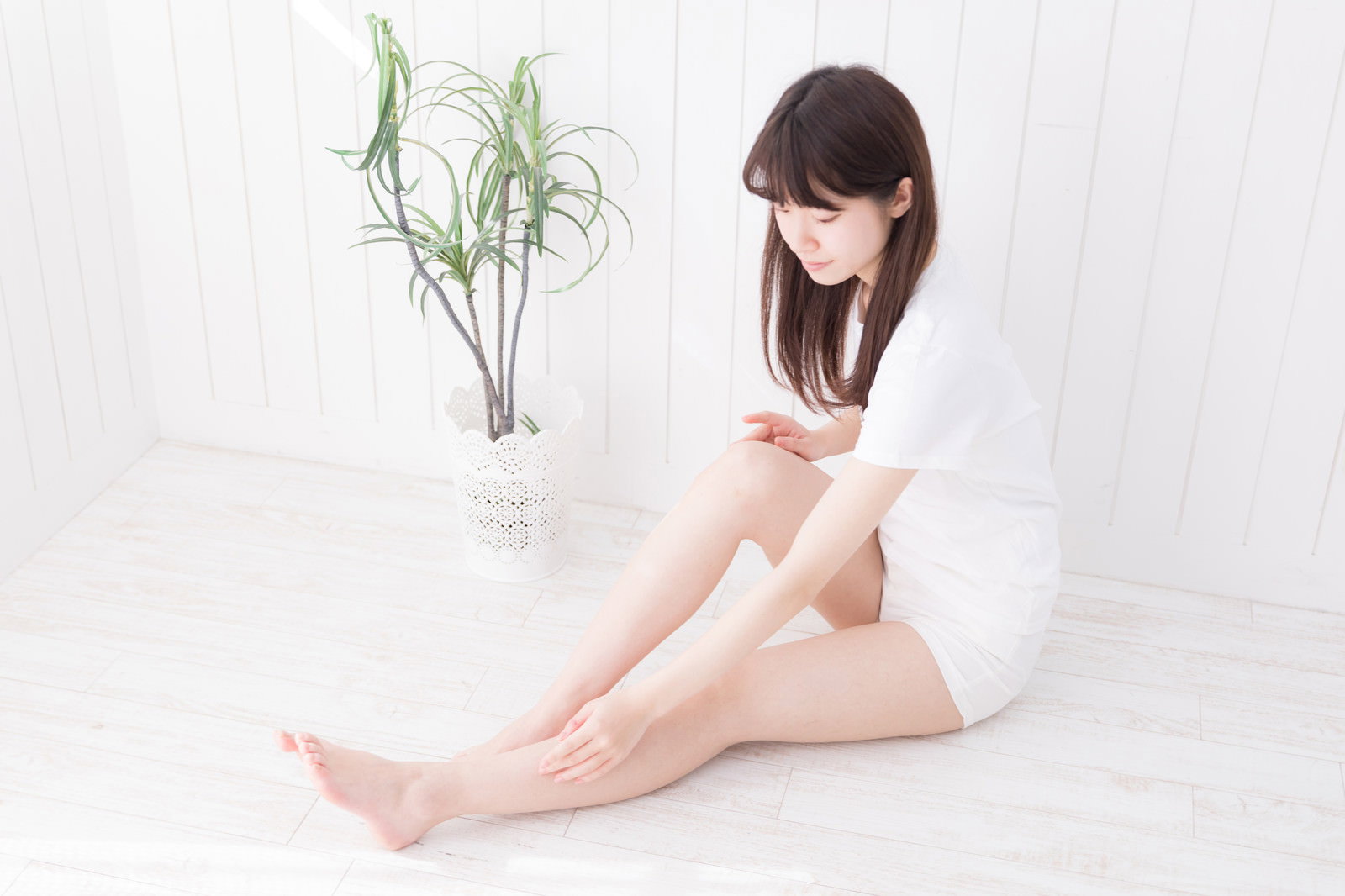 白い室内で白いワンピースを着た女性が床に座り、脚を伸ばして手入れされた素足を見せている［モデル：川子芹菜］