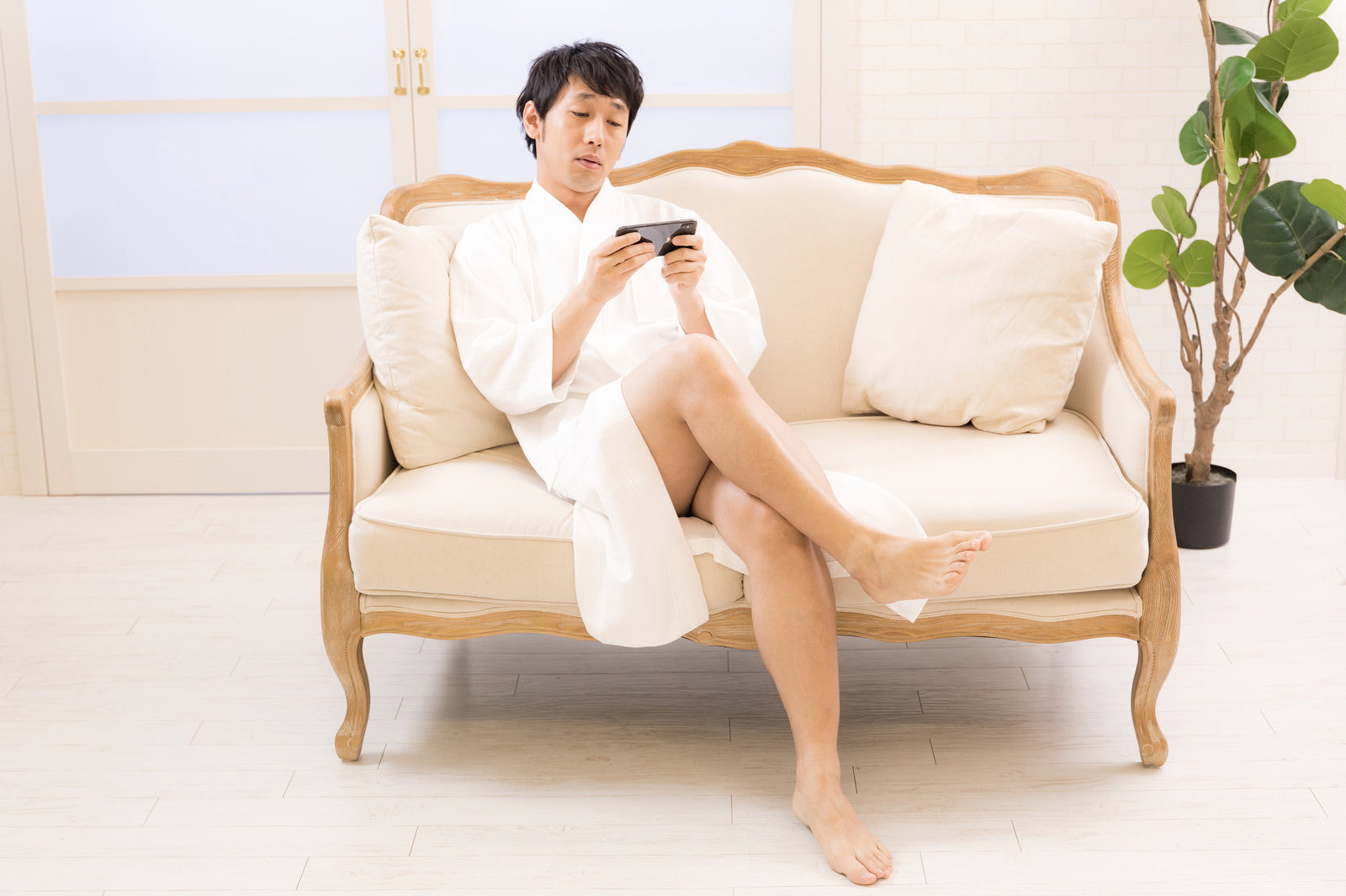 白いガウンを着てベージュのソファに座りスマホを見る男性［モデル：大川竜弥］