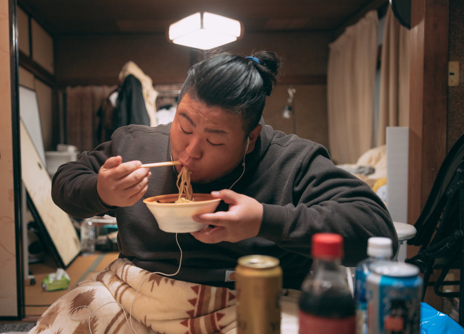 散らかった部屋でラーメンを食べるパーカー姿の肥満男性［モデル：ノリックス］