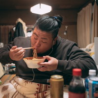散らかった部屋でラーメンをすする肥満男性の写真