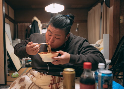 散らかった部屋でラーメンをすする肥満男性