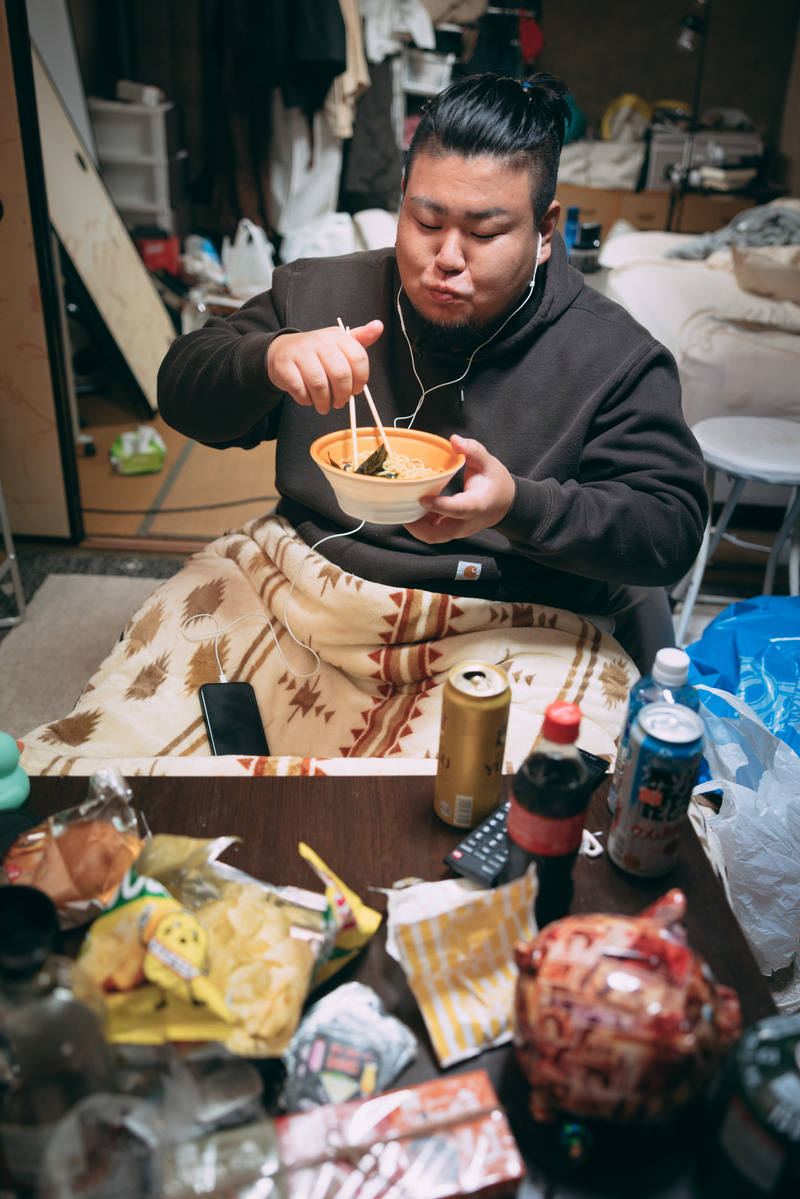 こたつに入ってラーメンを食べている黒パーカーの男性［モデル：ノリックス］