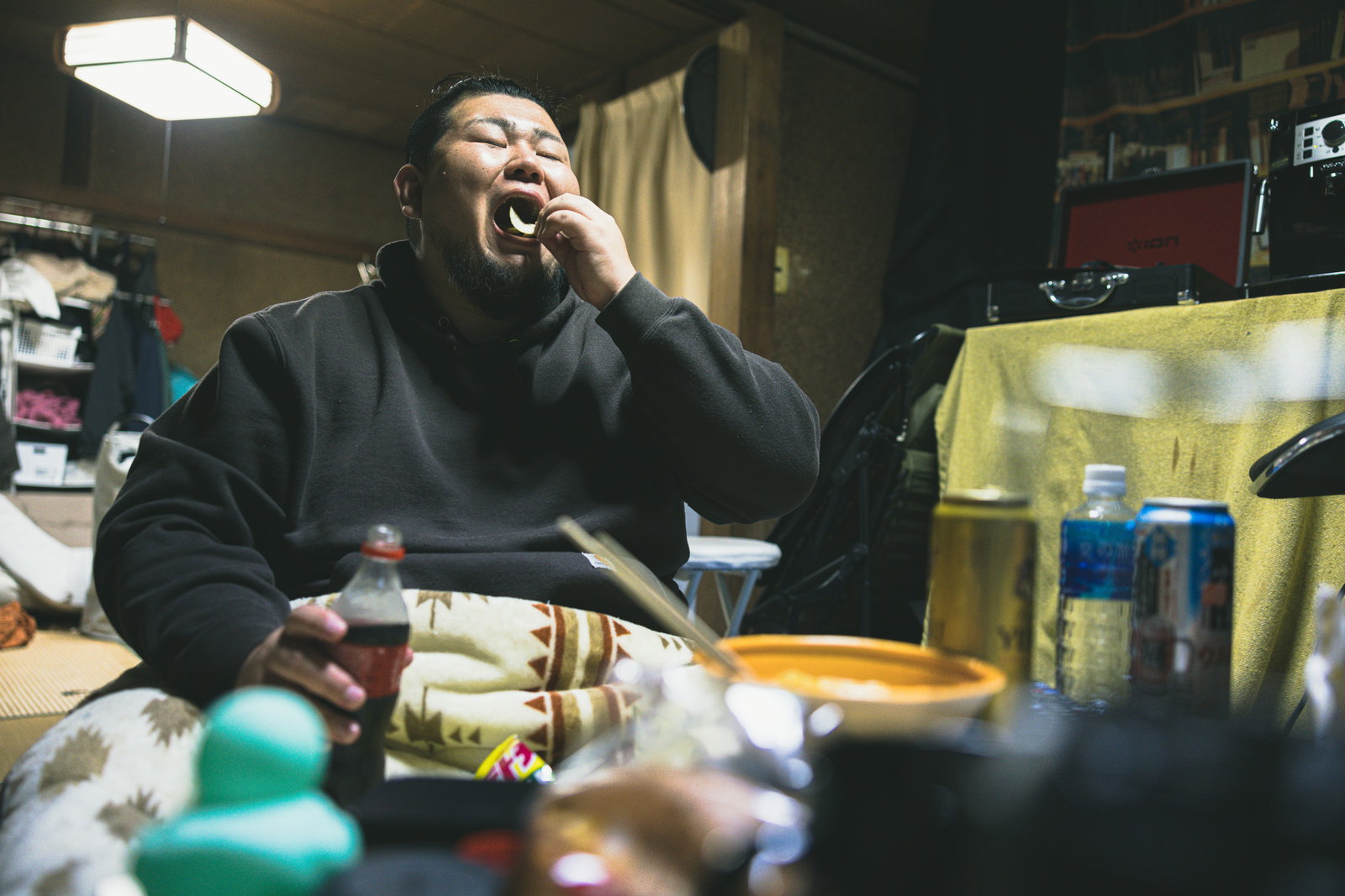 暗い部屋で黒いパーカーを着た無精髭の男性がお菓子を食べている様子［モデル：ノリックス］