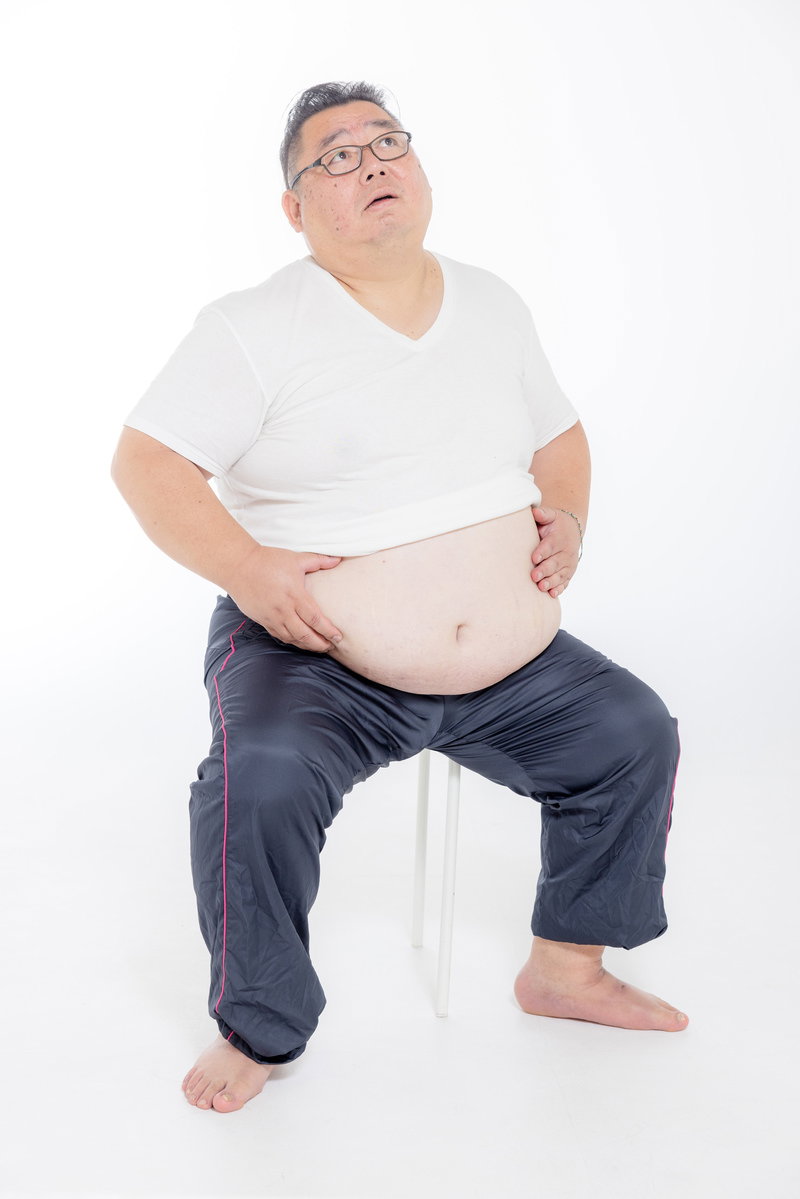 眼鏡をかけた男性が椅子に座り、お腹の脂肪に両手を当てている［モデル：ダルマ親方］