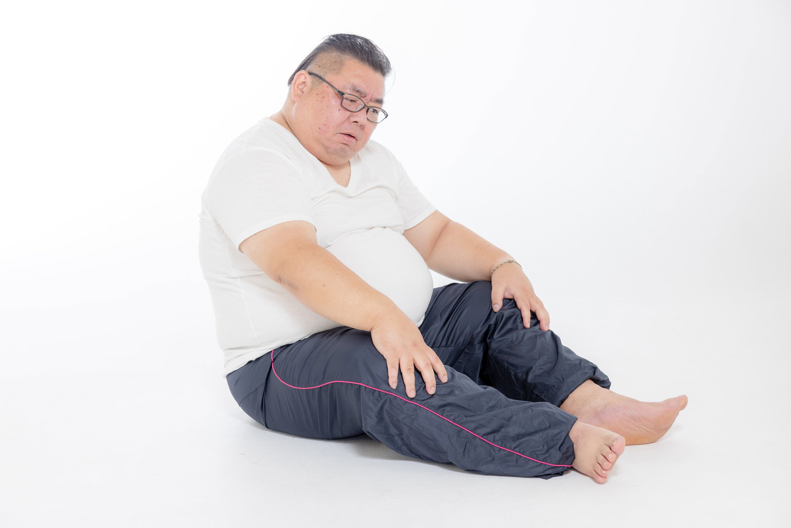 白いTシャツを着た太った男性が床に座り、お腹の脂肪のせいで姿勢が悪くなっている［モデル：ダルマ親方］