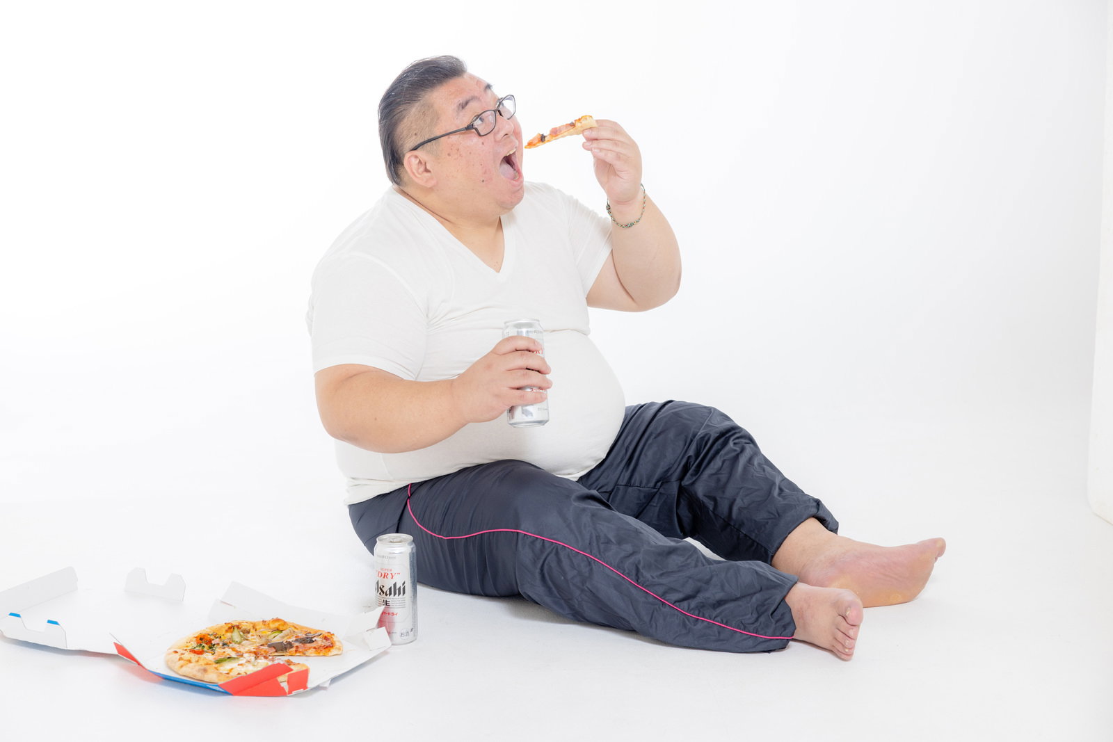 ビールを片手にピザを食べる膨よかな男性が足を伸ばしている［モデル：ダルマ親方］