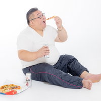 ビールを片手にピザを食べる膨よかな男性、肥満と飲酒の写真