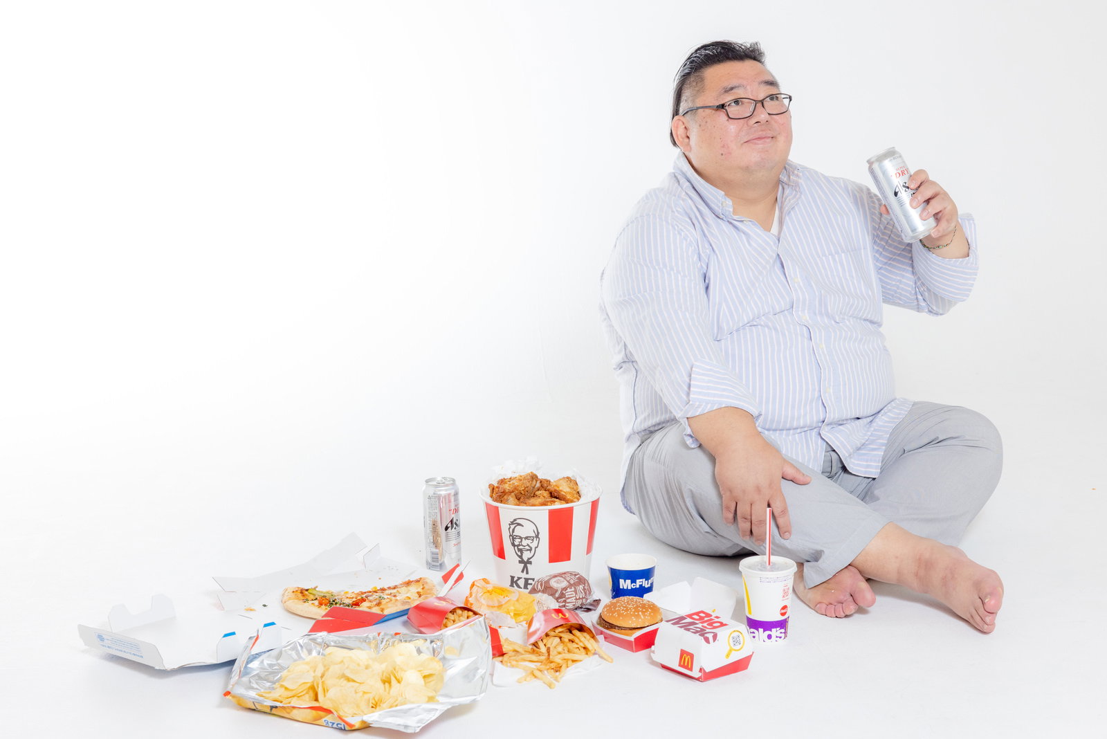 ジャンクフードに囲まれて胡坐をかき、スプレー缶を持つ膨よかな男性［モデル：ダルマ親方］