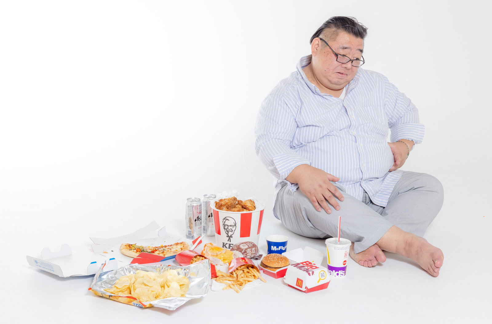 ジャンクフードに囲まれて胡坐をかき、腰のぜい肉を掴む眼鏡をかけた男性［モデル：ダルマ親方］