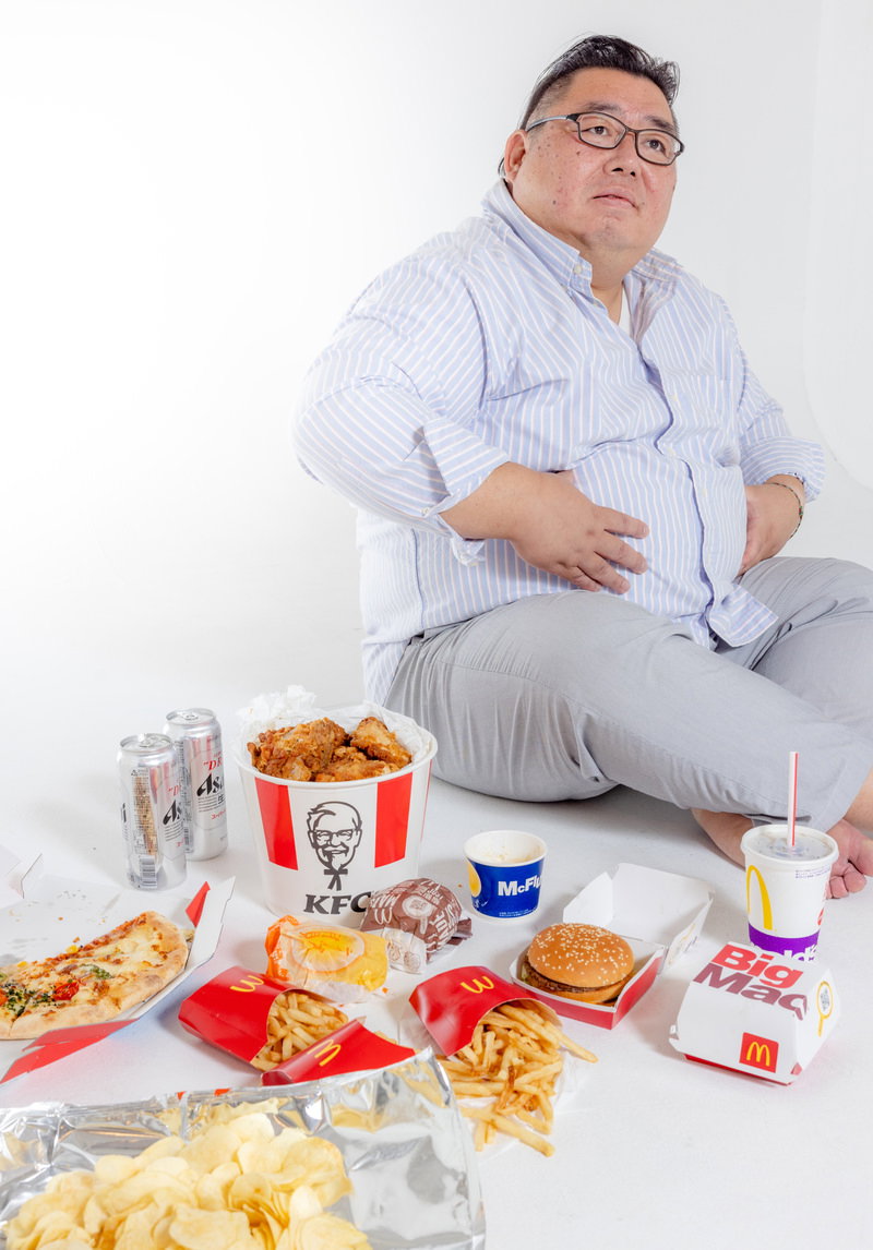 ファーストフードに囲まれて食べ過ぎた男性が床座りで腹部に手を当てている［モデル：ダルマ親方］