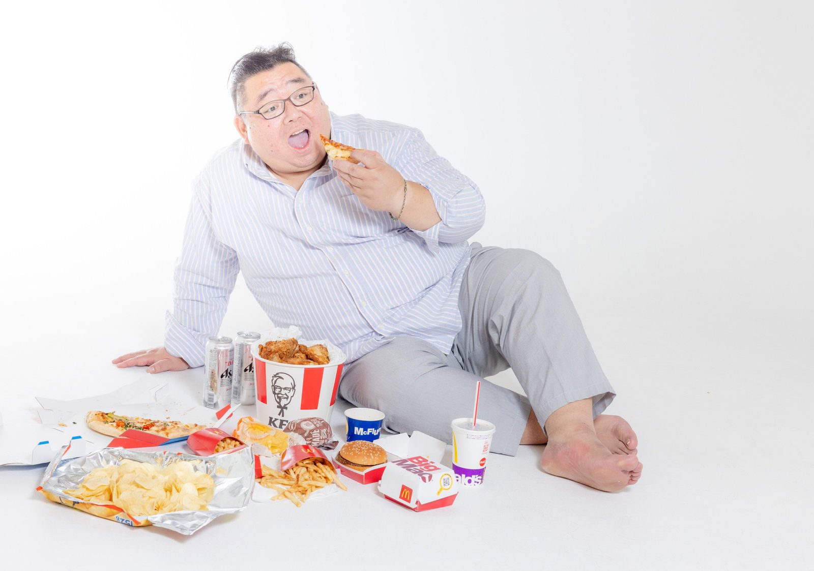 眼鏡をかけた男性がジャンクフードに囲まれてフライドチキンを食べている［モデル：ダルマ親方］