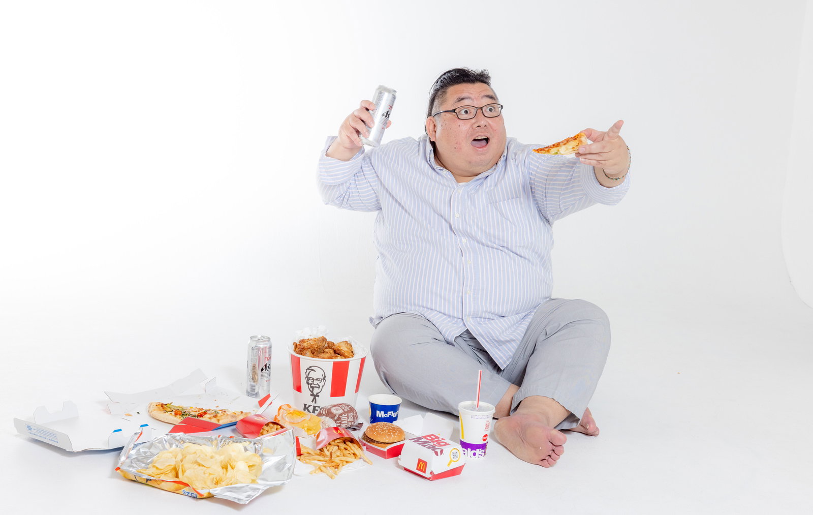 ジャンクフードとドリンクを持つ膨よかな男性がメガネをかけて笑顔で指を差している［モデル：ダルマ親方］