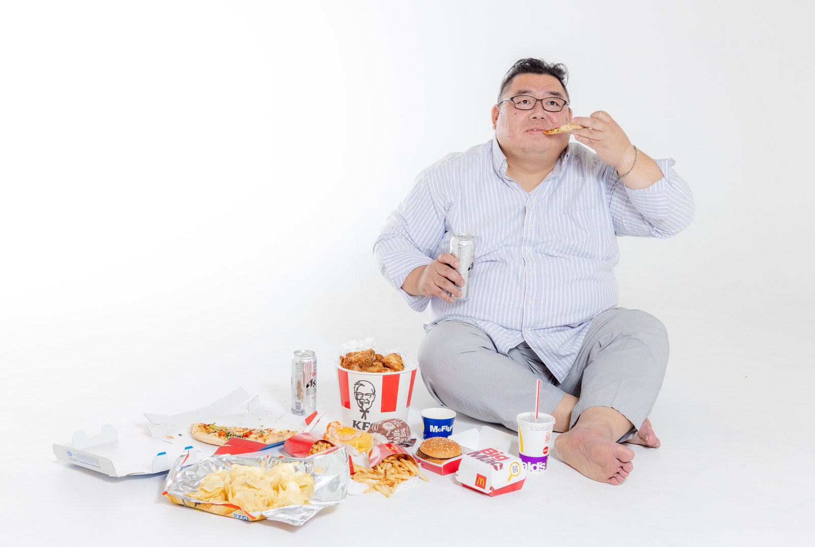ジャンクフードに囲まれて食べ物を食べる膨よかな男性［モデル：ダルマ親方］