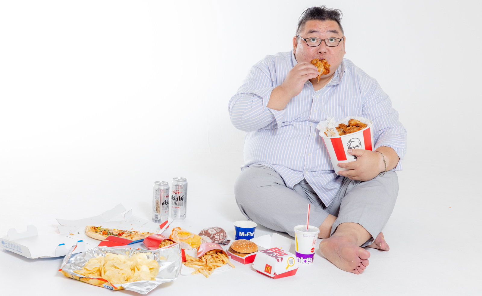 地べたに座ってファーストフードを食べるメガネをかけた太った男性［モデル：ダルマ親方］