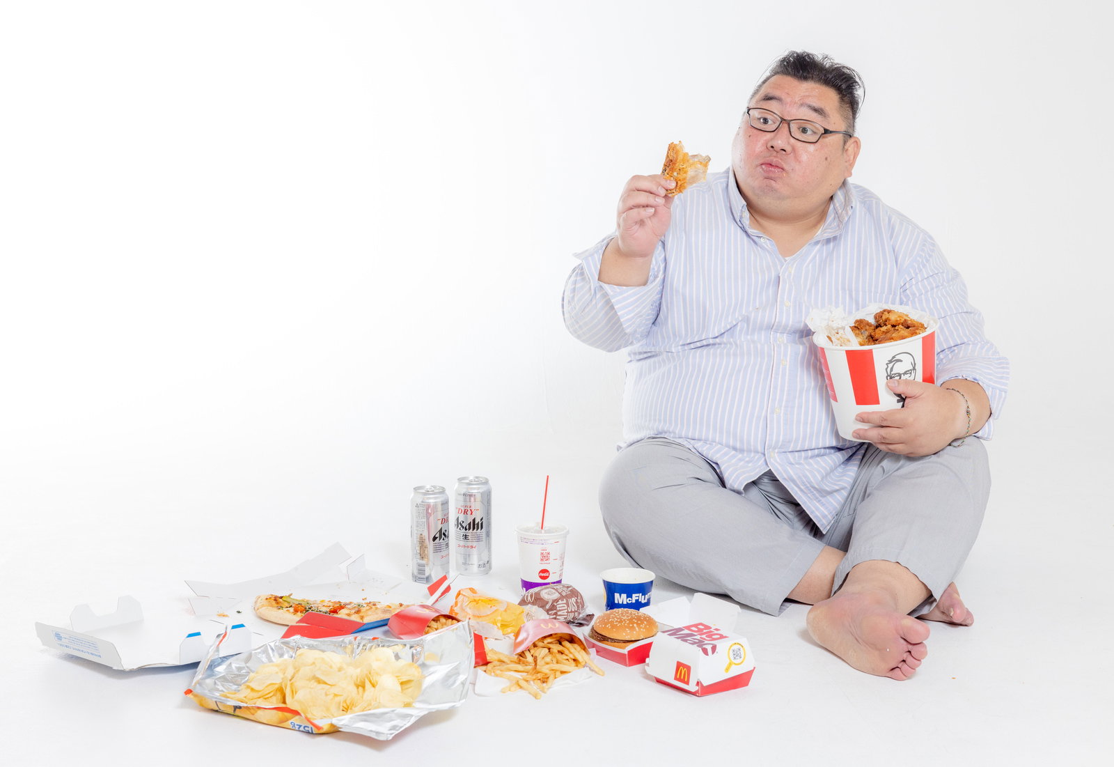 ジャンクフードに囲まれてフライドチキンを食べる眼鏡をかけた男性［モデル：ダルマ親方］