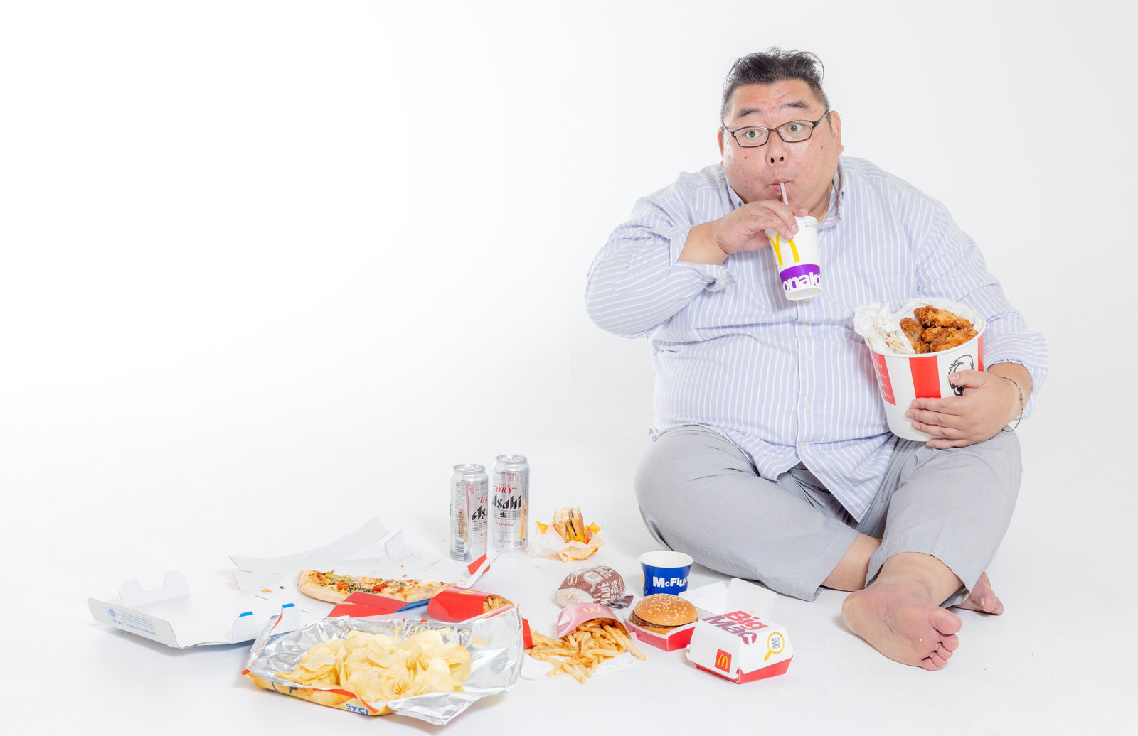 ファーストフードに囲まれながらドリンクを飲む眼鏡をかけた男性［モデル：ダルマ親方］