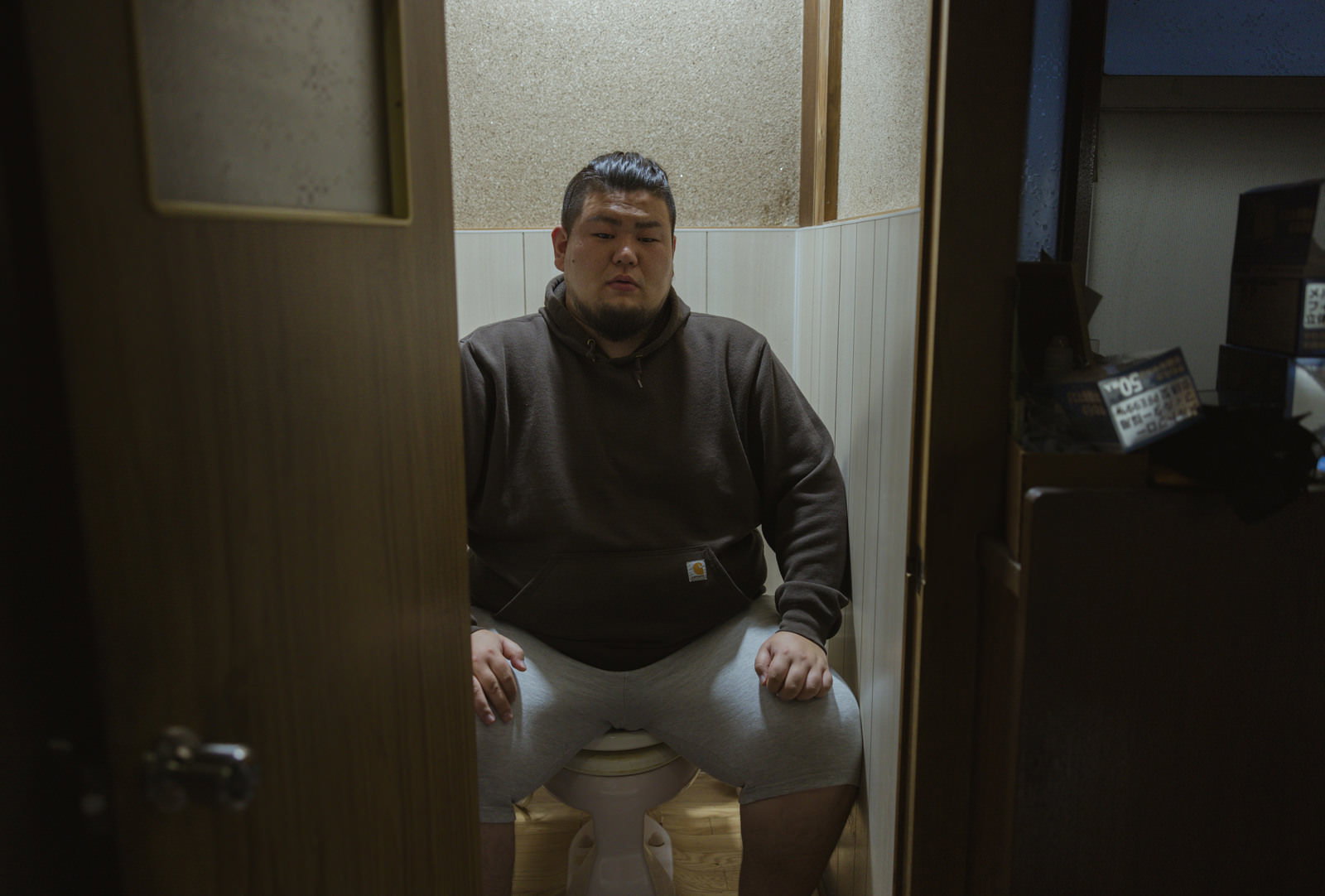 大柄な男性がトイレの便座にぼんやり座っている［モデル：ノリックス］