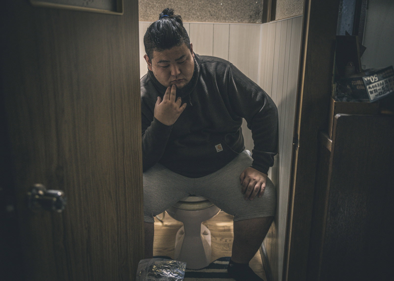 トイレの便座に座り、頭を抱える太った男性の写真［モデル：ノリックス］