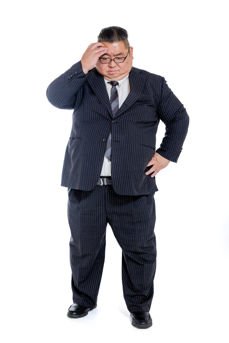 はち切れそうなスーツを着て困惑した表情で頭を掻く男性［モデル：ダルマ親方］