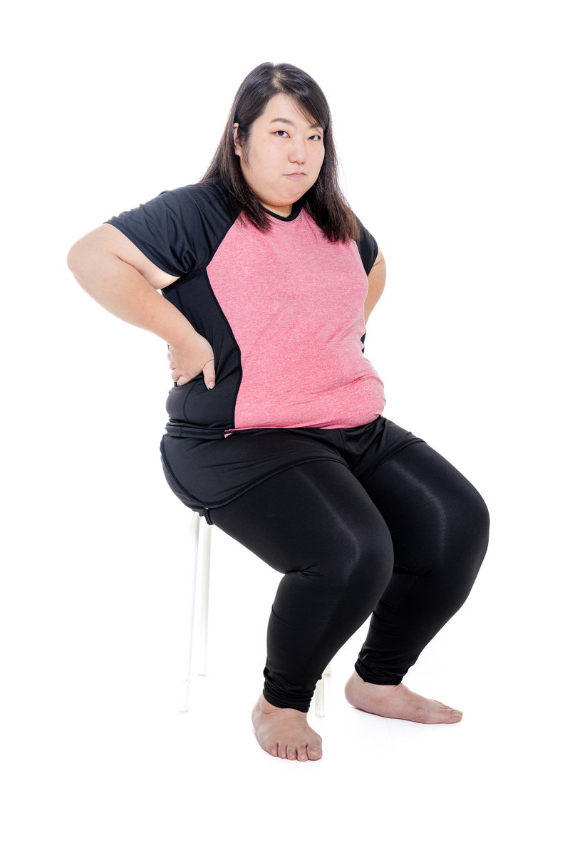 ピンク色のシャツで椅子に座り腰に手を当てるダイエット中の女性［モデル：KANA］