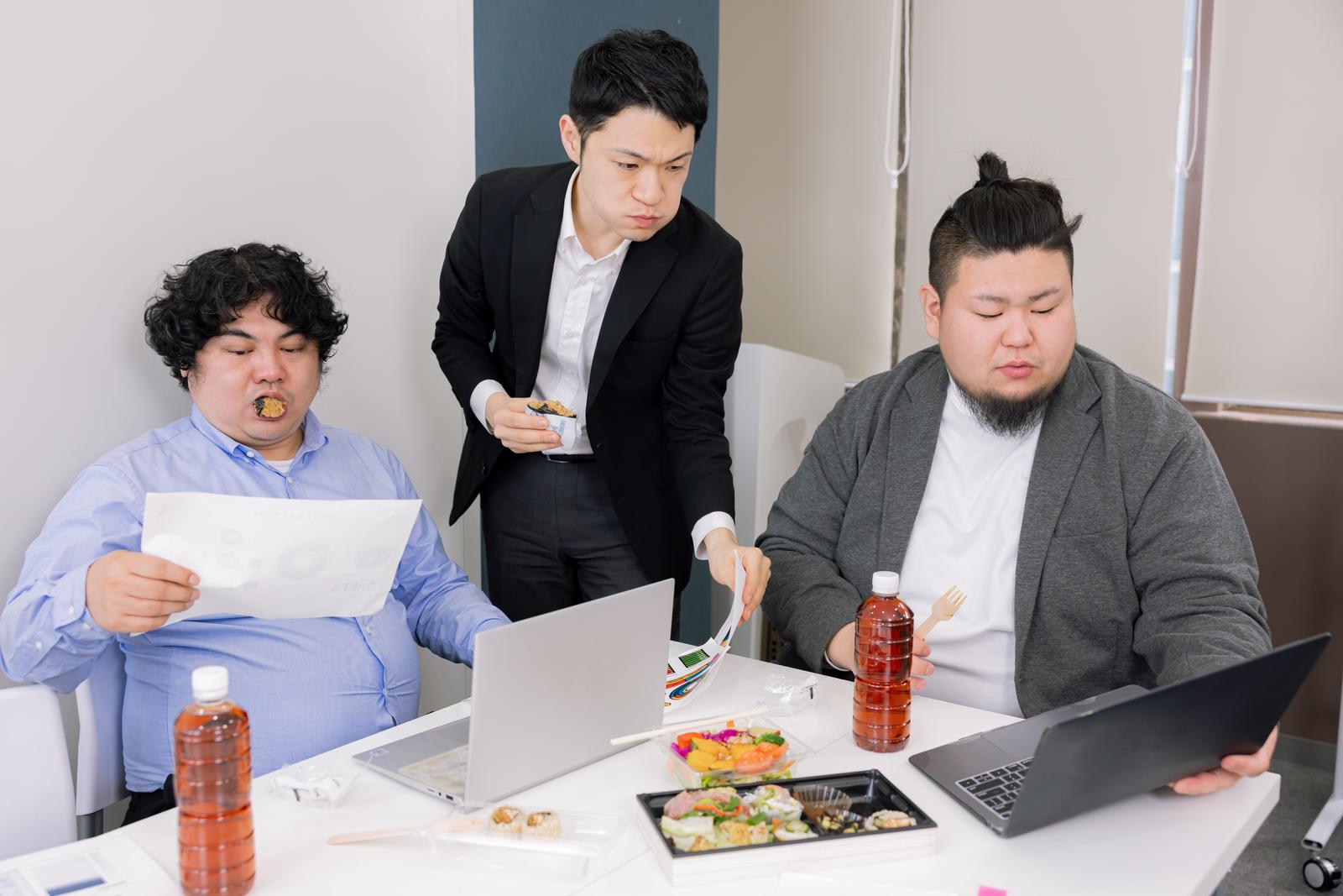 会議室でランチミーティング中に昼食を食べながら打ち合わせする男性たち［モデル：段田隼人 モデルリリース ノリックス］