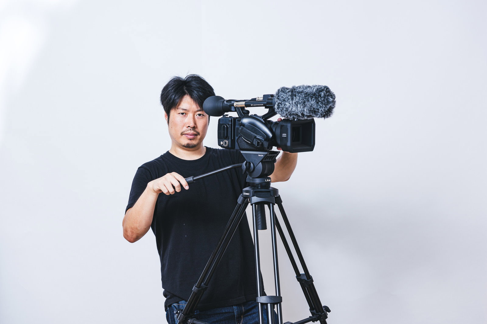 三脚に設置されたビデオカメラを操作して撮影するカメラマン［モデル：瀧野恵太］